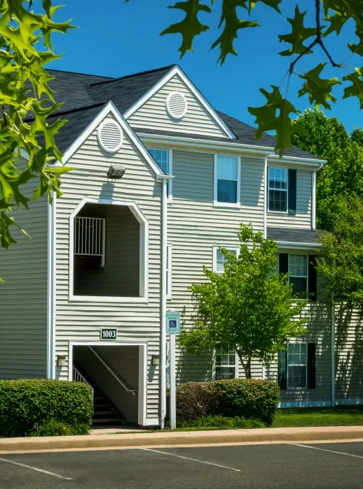 Three-story beige apartment building with white trim, surrounded by trees and greenery under a blue sky.