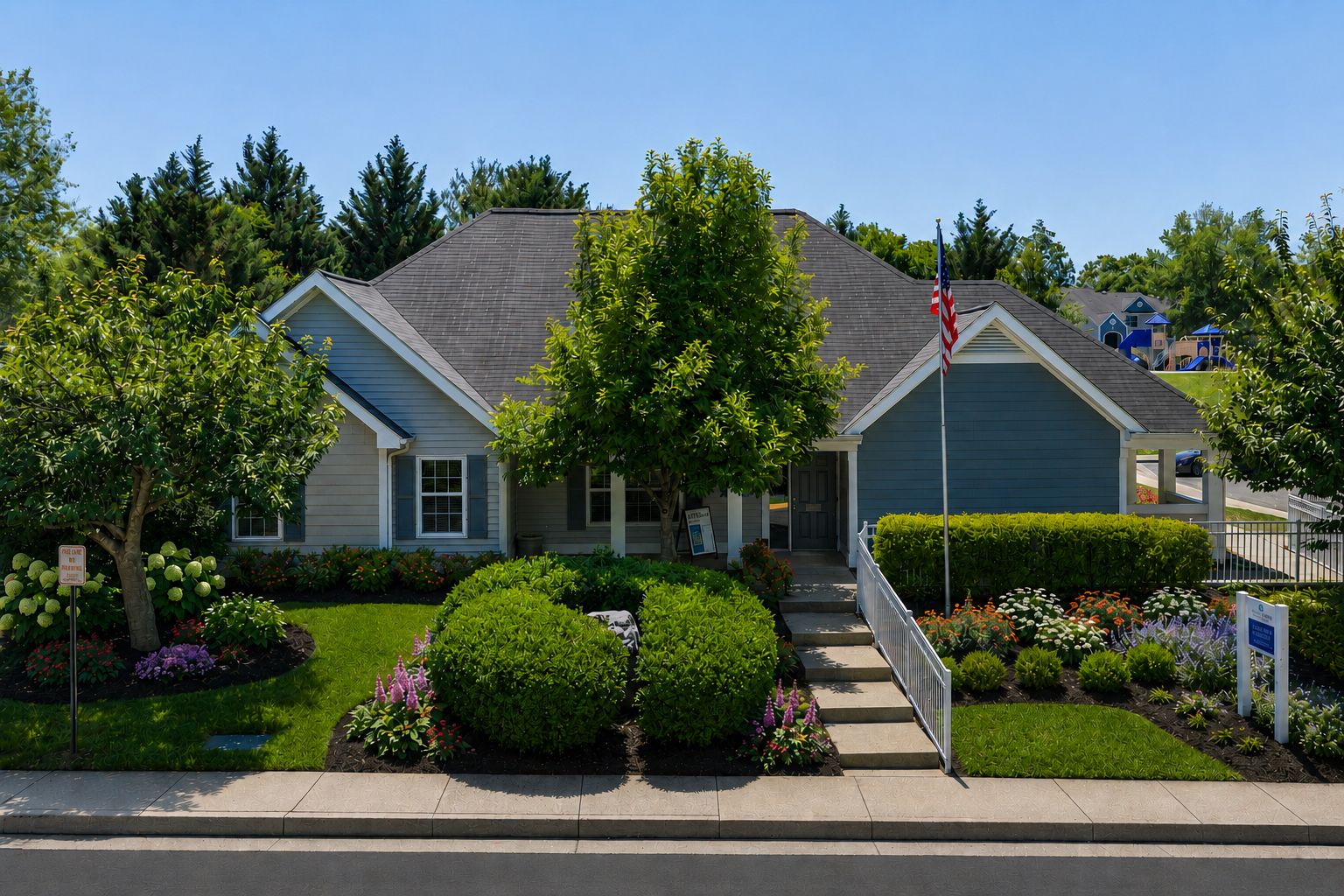 Blue suburban house with a front garden, trees, a flagpole, and a playground in the background on a sunny day.