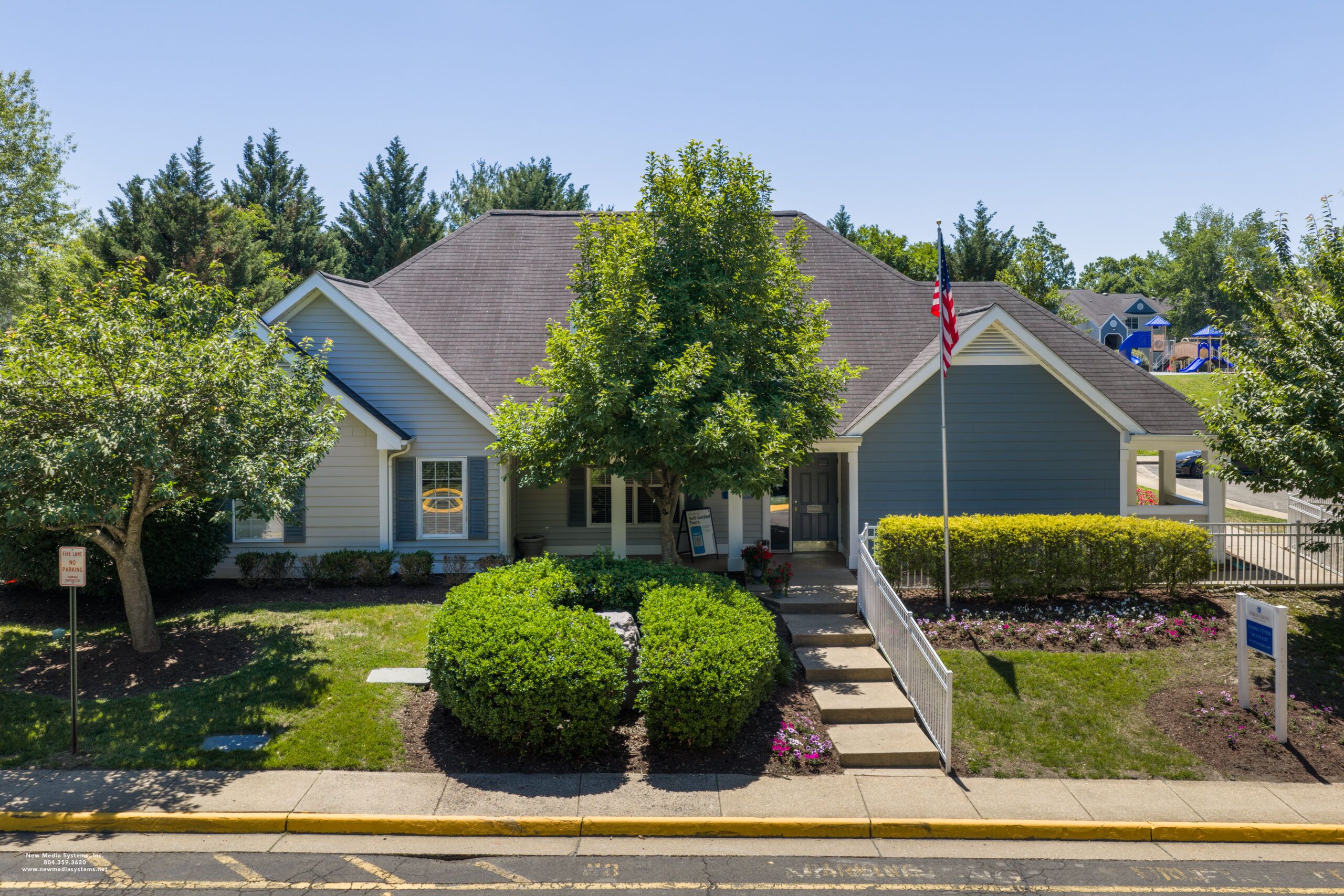 Blue-gray house with a front porch, flagpole, trees, and landscaped bushes on a sunny day.