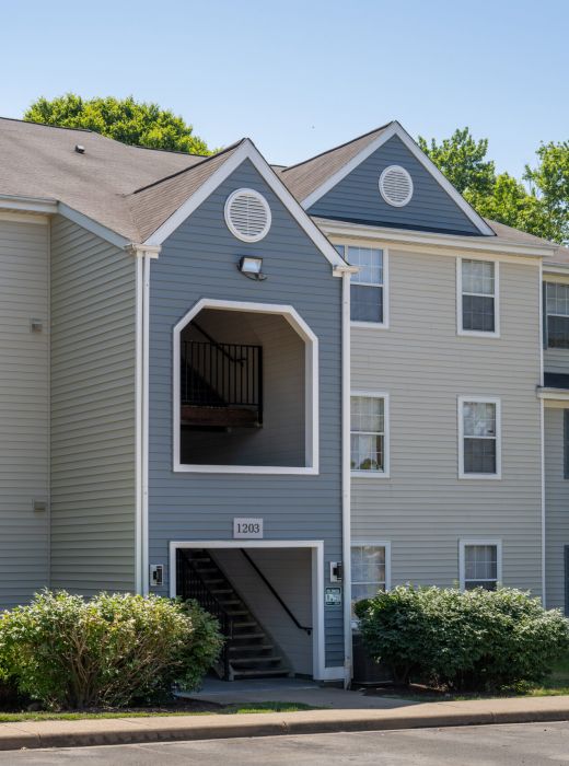 Three-story beige apartment building with an open stairwell entrance and surrounded by trees and bushes.