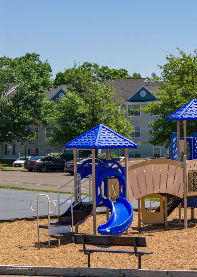 Playground with blue slides, climbing structures, and swings on a sunny day, with trees and apartments in background.