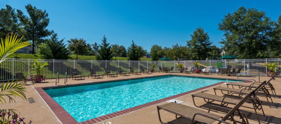 Outdoor swimming pool with lounge chairs, surrounded by a fence and trees under a clear blue sky.