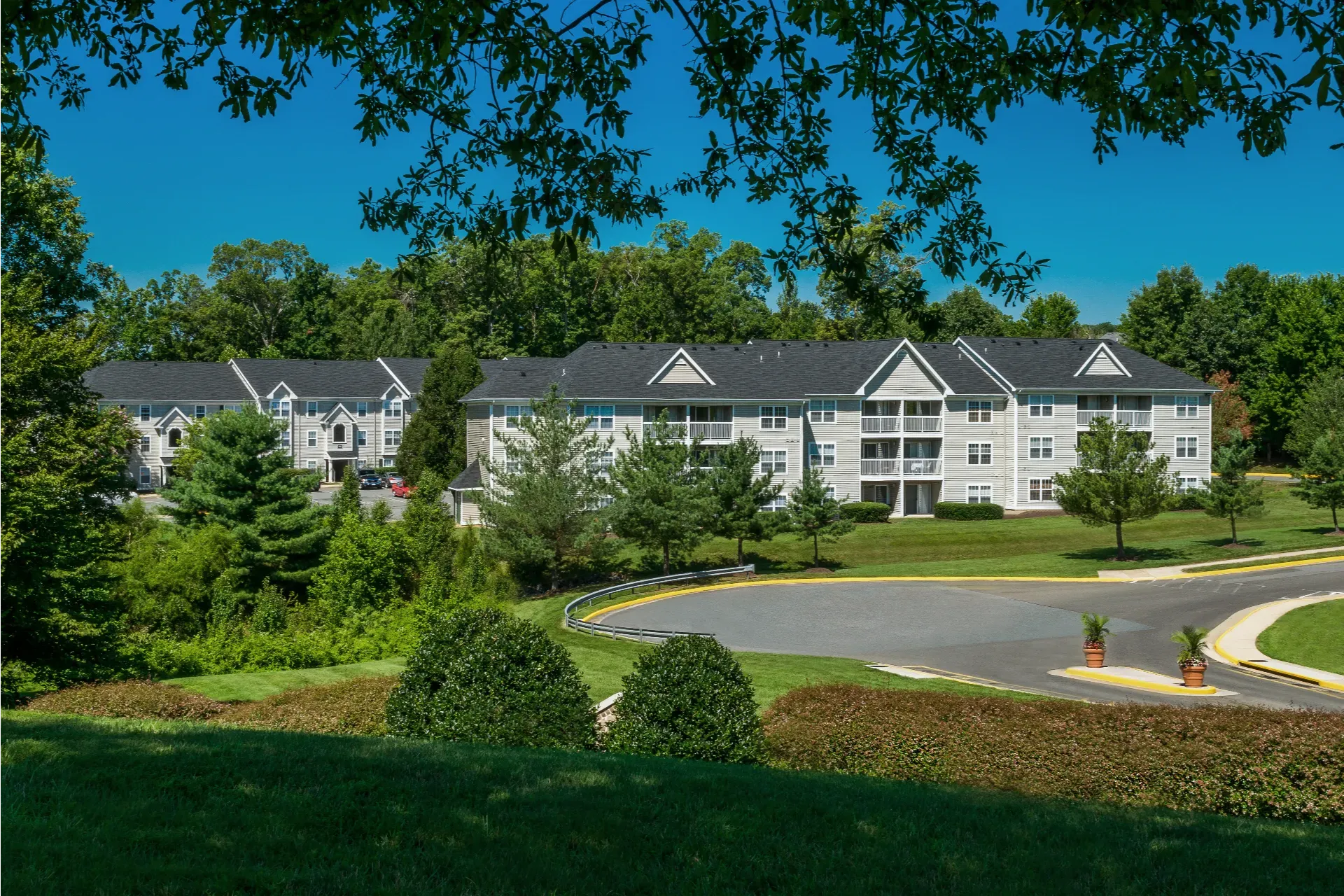 Three-story apartment buildings with balconies, surrounded by green trees and a curved driveway under a blue sky.