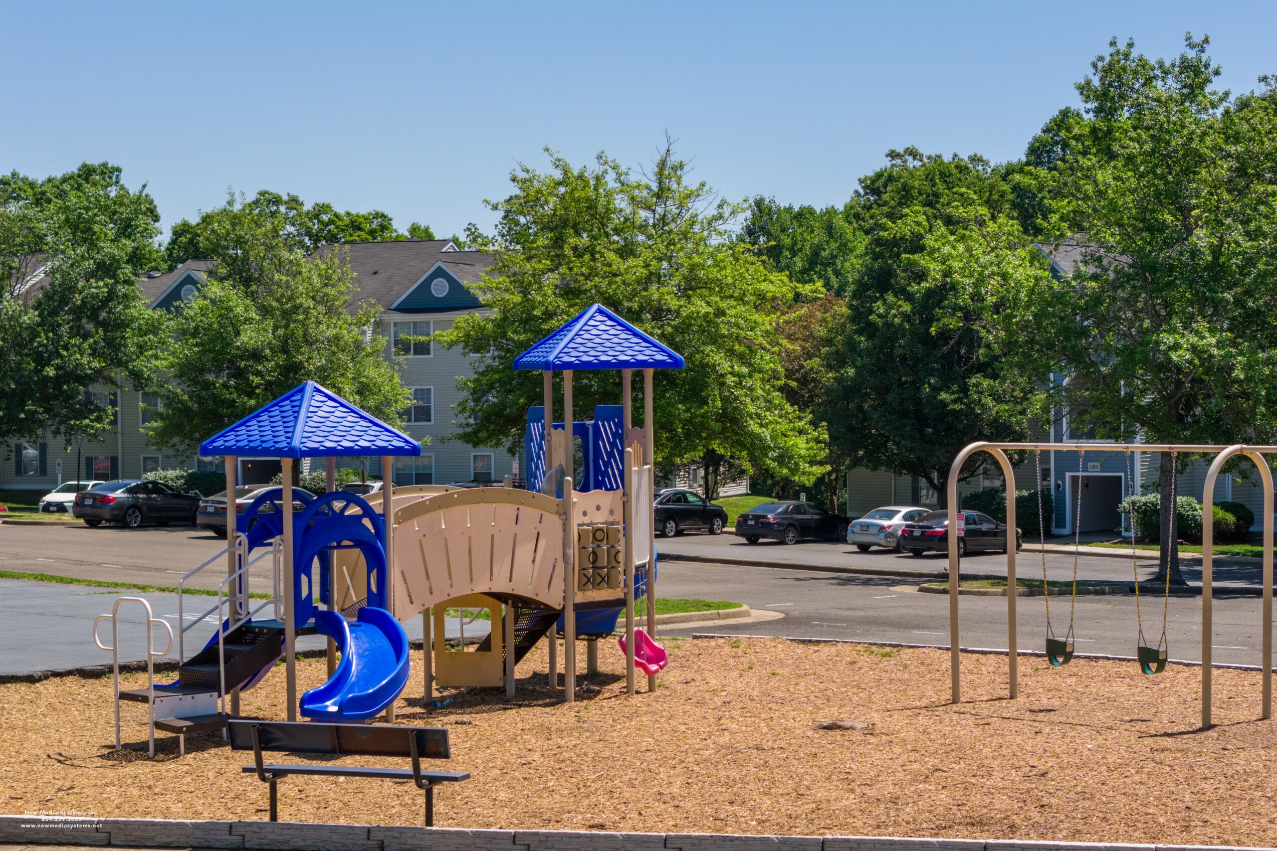 Playground with blue slides, climbing structures, and swings on a sunny day, with trees and apartments in background.