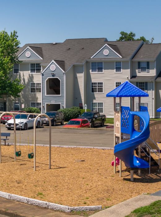 Playground with blue slides and swings in front of an apartment building and parked cars on a sunny day.