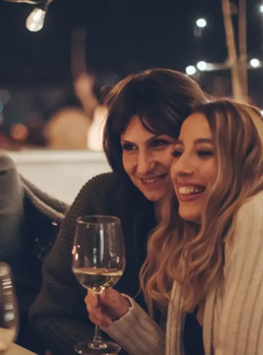 Three women smiling and enjoying wine together at a cozy, string-lit outdoor gathering at night.